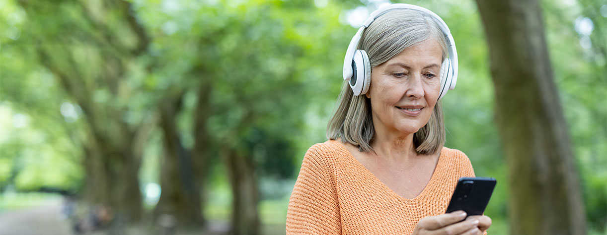 Mature woman wearing headphones using smartphone in park. 