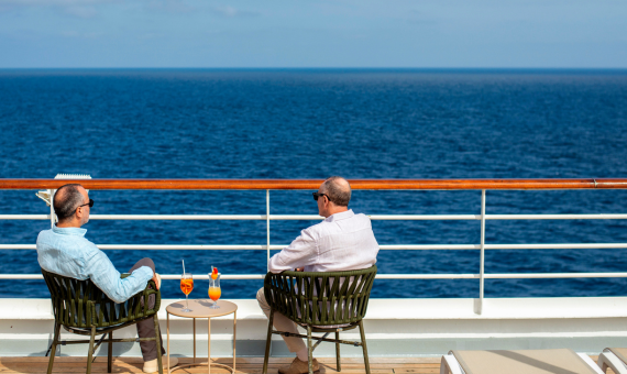 Two guests relax in chairs on a ship's deck, sharing orange cocktails as they look out over a calm blue sea.